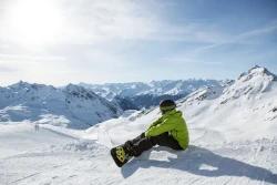 Snowboarder sitzt mit angeschnalltem Snowboard auf der Piste un schaut ins Tal